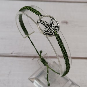 May include: A green braided bracelet with a silver charm featuring a fern design.
