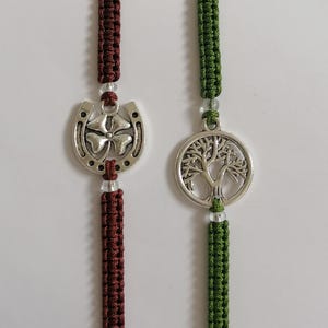 May include: Two macrame bracelets. One is burgundy with a horseshoe and four-leaf clover charm. The other is green with a tree of life charm. Both bracelets have clear beads and are on a white background.