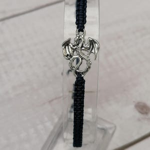 May include: A dark blue macrame bracelet with a silver dragon charm. The dragon charm has detailed wings and a serpentine body. The bracelet is displayed on a clear acrylic stand, highlighting the macrame knotting.