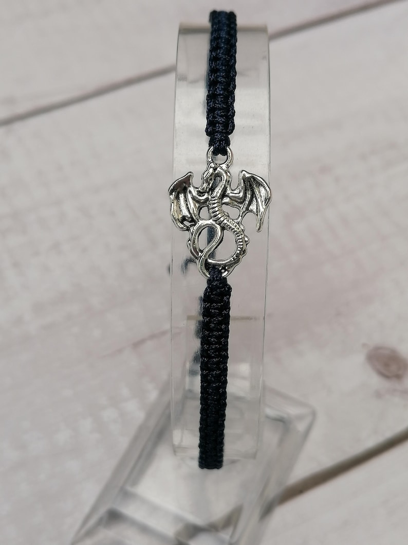 May include: A dark blue macrame bracelet with a silver dragon charm. The dragon charm has detailed wings and a serpentine body. The bracelet is displayed on a clear acrylic stand, highlighting the macrame knotting.