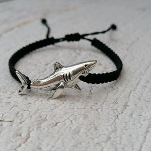 May include: A black braided cord bracelet with a silver shark charm. The bracelet is tied with a knot and has a silver shark charm.