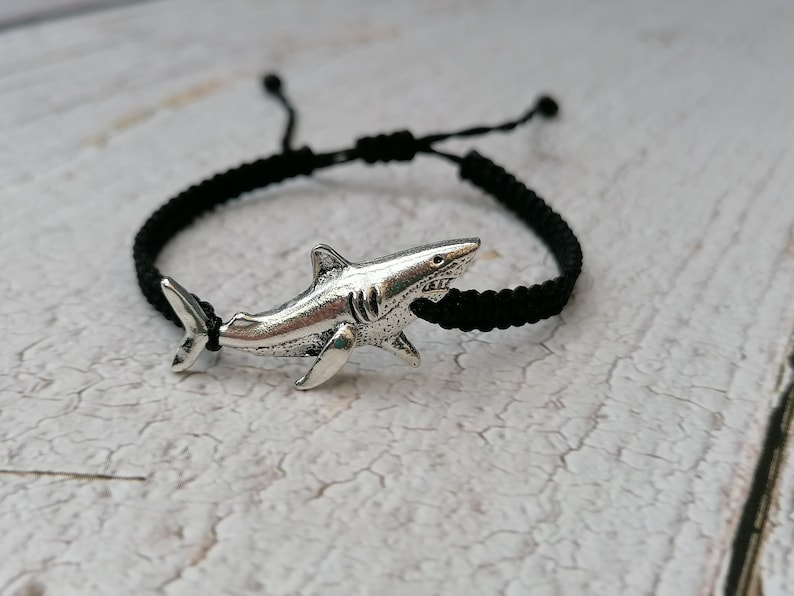 May include: A black braided cord bracelet with a silver shark charm. The bracelet is tied with a knot and has a silver shark charm.