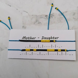 Puede incluir: Dos pulseras de amistad con cuentas negras y amarillas en una tarjeta blanca con el texto "Madre - Hija".