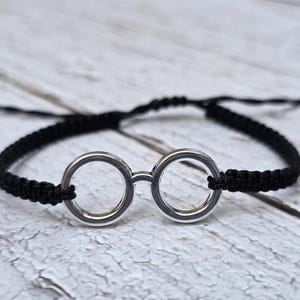 Peut inclure: Un bracelet macramé noir avec un charm en forme de lunettes de couleur argentée. Le bracelet réglable est présenté sur une surface en bois blanc. Le charm de lunettes a deux verres circulaires reliés par un pont.