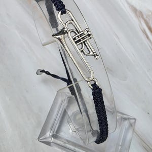 May include: A dark navy blue braided cord bracelet featuring a silver-toned metal trumpet charm. The bracelet has an adjustable sliding knot closure and is displayed on a clear curved stand to showcase the instrument design.