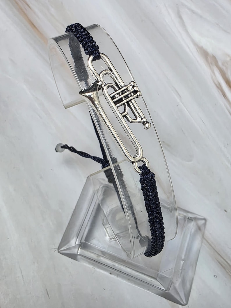May include: A dark navy blue braided cord bracelet featuring a silver-toned metal trumpet charm. The bracelet has an adjustable sliding knot closure and is displayed on a clear curved stand to showcase the instrument design.