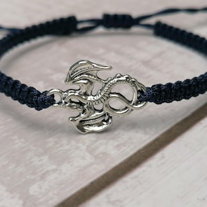 May include: A navy blue macrame bracelet with a silver-coloured dragon charm. The adjustable bracelet is made of woven cord and has a sliding knot closure. The dragon charm is detailed with wings and scales.