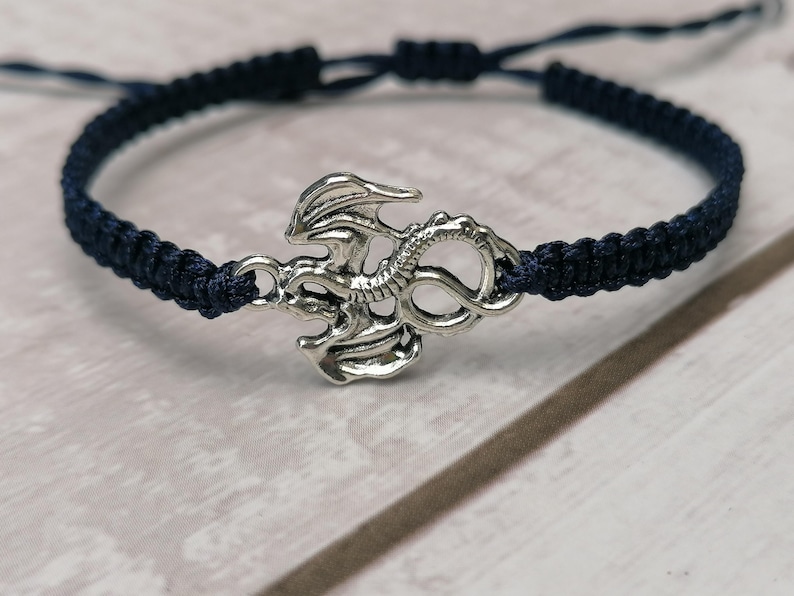 May include: A navy blue macrame bracelet with a silver-coloured dragon charm. The adjustable bracelet is made of woven cord and has a sliding knot closure. The dragon charm is detailed with wings and scales.
