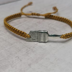 Puede incluir: Un brazalete trenzado dorado con un dije de pincel plateado. El brazalete es ajustable y puede ser usado por cualquier persona.