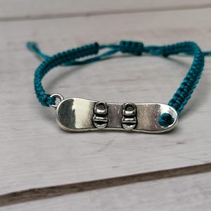 Könnte beinhalten: Armband aus geflochtenem türkisfarbenem Band mit einem silbernen Anhänger in Form eines Snowboards.