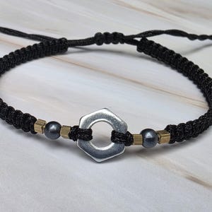 May include: A black cord bracelet featuring a central silver-toned hexagonal metal nut. The design includes small metallic cube spacers and dark grey spherical beads on either side of the nut, finished with an adjustable braided cord closure.