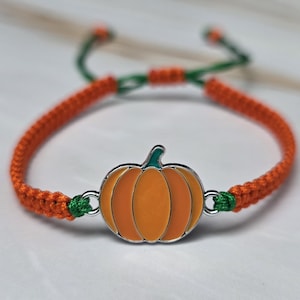 May include: A bracelet featuring a central metal pumpkin charm with orange enamel fill and a green stem. The band is constructed from braided orange cord with green cord accents at the connection points and adjustable sliding knots at the back.