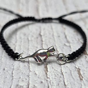 May include: A black braided cord bracelet with a silver cat charm and a ball of yarn. The bracelet has an adjustable sliding knot. The cat charm is detailed. The bracelet is set against a light-coloured, textured background.