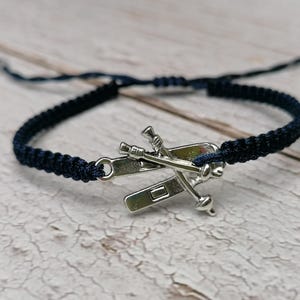 Könnte beinhalten: Ein marineblaues Makramee-Armband mit einem silberfarbenen Anhänger mit einem Boot und gekreuzten Rudern. Das Armband ist verstellbar und liegt auf einer hellen Holzoberfläche.