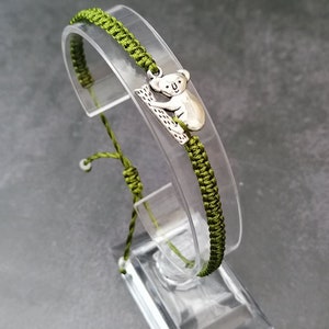 May include: A green braided bracelet with a silver koala charm. The bracelet is displayed on a clear plastic stand.