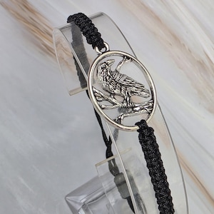May include: A black braided cord bracelet featuring a circular silver-toned metal charm. The charm depicts a detailed raven perched on a branch within an oval frame. The bracelet has a woven texture and adjustable cord ends for a secure fit.