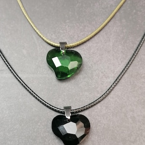 Puede incluir: Dos collares con colgantes en forma de corazón. Un colgante es de cristal verde esmeralda facetado, y el otro es de cristal negro facetado. Ambos colgantes están suspendidos de una fianza plateada en un collar de cordón. El collar superior tiene un cordón de color beige claro, y el inferior tiene un cordón negro.