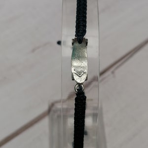Puede incluir: Un brazalete trenzado negro con un cierre plateado. El cierre tiene forma de un peque&ntilde;o rect&aacute;ngulo plano con bordes redondeados.