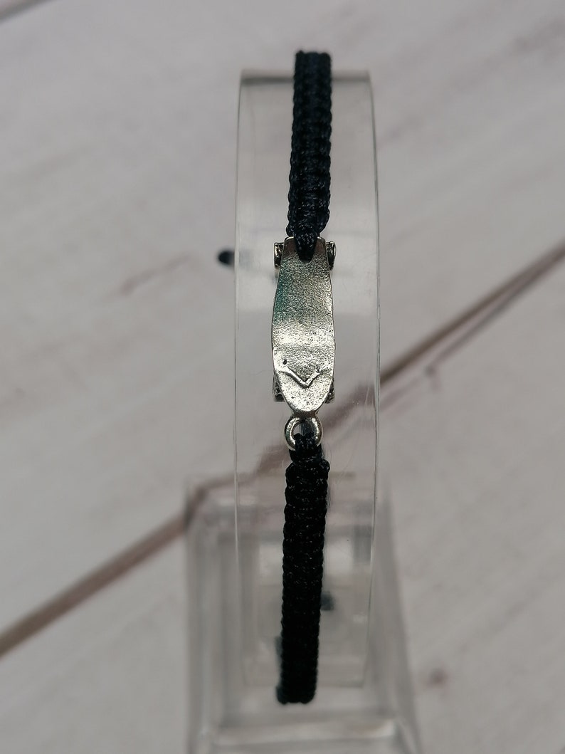 Puede incluir: Un brazalete trenzado negro con un cierre plateado. El cierre tiene forma de un peque&ntilde;o rect&aacute;ngulo plano con bordes redondeados.