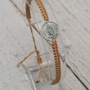 Può includere: Un braccialetto in macramè beige con un ciondolo circolare color argento raffigurante mani in preghiera. Il braccialetto è regolabile con nodi scorrevoli. Il cordoncino in macramè è marrone chiaro.
