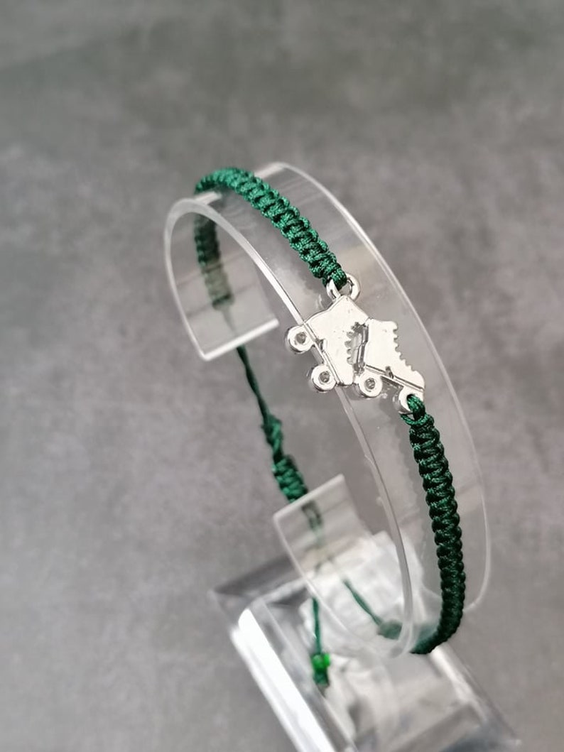 May include: A dark green braided cord bracelet featuring a silver-toned metal charm shaped like a pair of roller skates. The bracelet is constructed with a woven knot pattern and includes adjustable cord ends for a secure fit on the wrist.