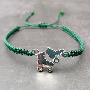 May include: A dark green woven cord bracelet featuring a silver-toned metal charm shaped like a pair of roller skates. The bracelet includes an adjustable sliding knot closure with small green beads at the ends of the cord strings.