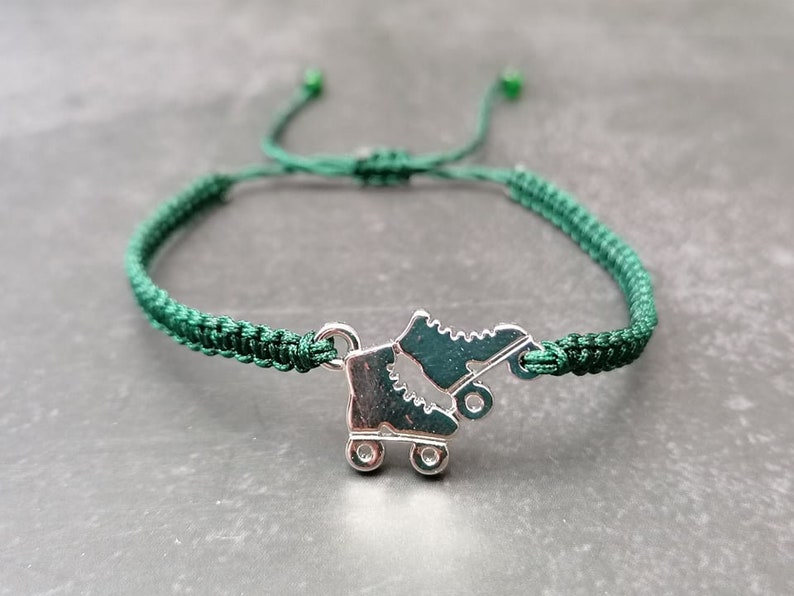 May include: A dark green woven cord bracelet featuring a silver-toned metal charm shaped like a pair of roller skates. The bracelet includes an adjustable sliding knot closure with small green beads at the ends of the cord strings.