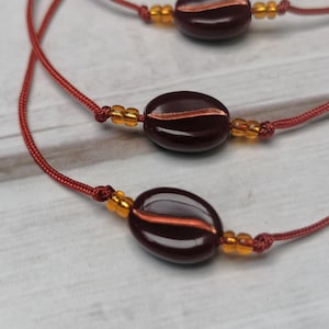 Könnte beinhalten: Drei rote Schnüre mit braunen Kaffeebohnen-förmigen Perlen und kleinen goldenen Perlen.