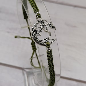 Pode incluir: Um bracelete trançado verde com um amuleto de prata de uma árvore crescendo dentro de uma lua crescente.