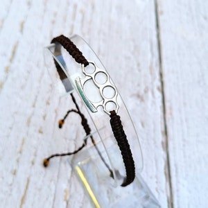 May include: A brown braided bracelet with a silver metal charm in the shape of a knuckle duster.