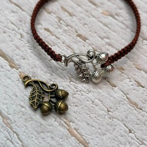 Könnte beinhalten: Ein braunes Kordelarmband mit einem silbernen Eichel-Anhänger. Der Anhänger zeigt ein Blatt und drei Eicheln.