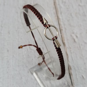 May include: A brown braided bracelet with a silver arrow charm. The arrow is in a circle and has a feather detail.