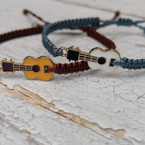Könnte beinhalten: Zwei Freundschaftsarmbänder mit Gitarrenanhängern. Ein Armband ist braun und das andere blau. Die Gitarrenanhänger sind gelb und weiß.