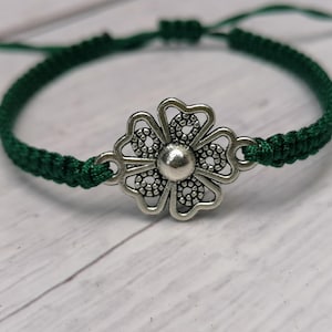 May include: A green braided bracelet with a silver flower charm. The flower charm has a detailed design with a circular center.