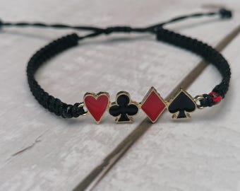Puede incluir: Una pulsera de cordón negro con cuatro dijes metálicos en forma de palos de baraja: un corazón rojo, un trébol negro, un diamante rojo y una pica negra. Los dijes están dispuestos en fila sobre un cordón trenzado negro con lazos ajustables.