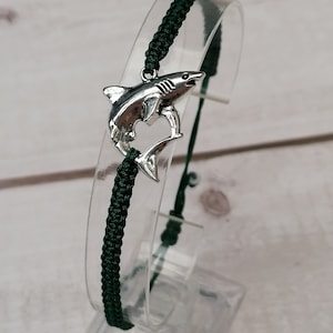 Puede incluir: Un brazalete ajustable de color verde oscuro con un dije de tiburón plateado.