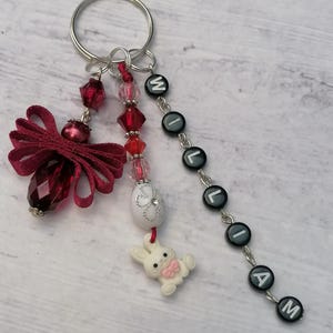 Puede incluir: Un llavero de temática de Pascua rojo y blanco con un amuleto de conejito blanco, un amuleto de huevo blanco con purpurina plateada y un lazo rojo. El llavero también presenta una cadena plateada con cuentas negras que deletrean el nombre "Liam".