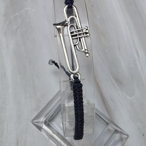 May include: A dark navy blue cord bracelet featuring a silver-toned metal trumpet charm. The bracelet is constructed with a woven cord design and includes a central musical instrument charm that is securely attached to the cord structure.