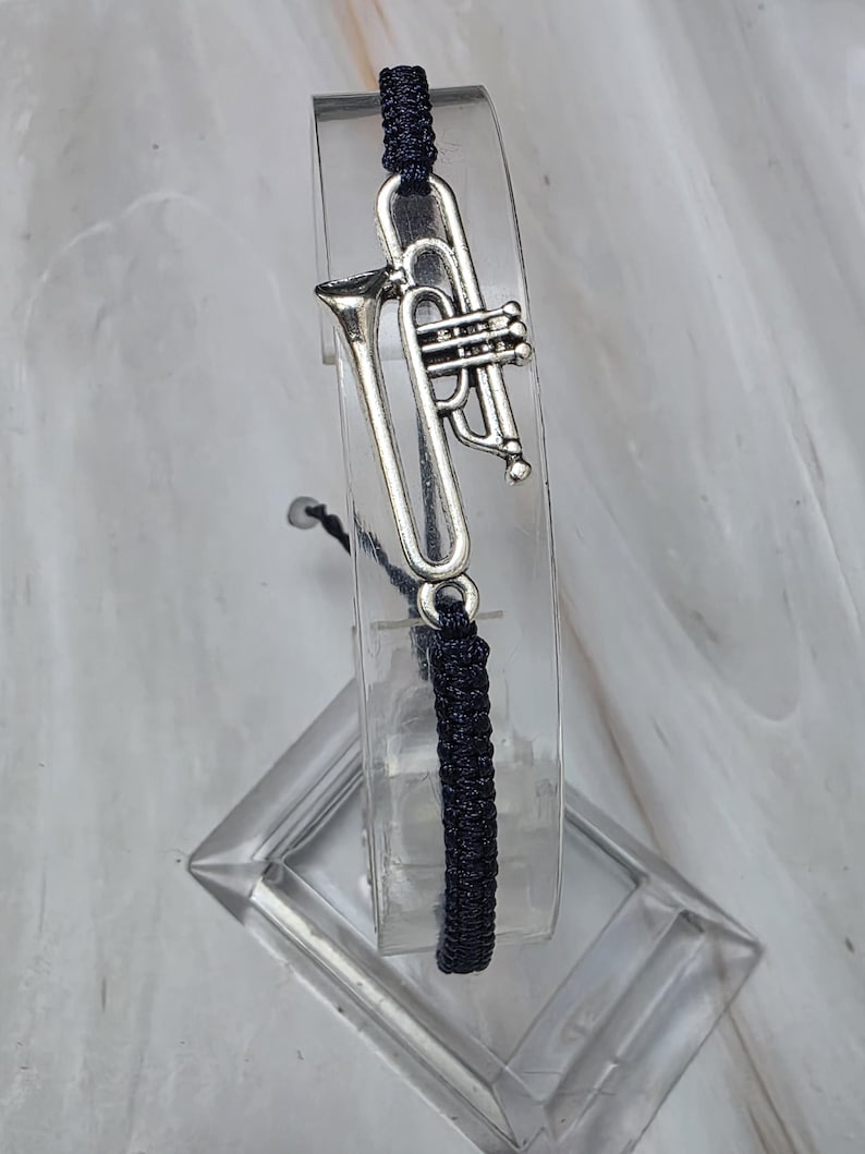 May include: A dark navy blue cord bracelet featuring a silver-toned metal trumpet charm. The bracelet is constructed with a woven cord design and includes a central musical instrument charm that is securely attached to the cord structure.