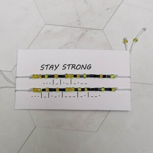 Può includere: Due braccialetti dell'amicizia con perline nere e verdi su una carta bianca con il testo "STAY STRONG".
