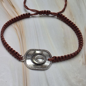 May include: A brown macrame bracelet featuring a silver-colored cowboy hat charm. The adjustable bracelet has a woven design and a sliding knot closure. The hat charm has a detailed design with a central oval accent.