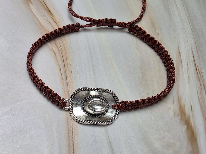 May include: A brown macrame bracelet featuring a silver-colored cowboy hat charm. The adjustable bracelet has a woven design and a sliding knot closure. The hat charm has a detailed design with a central oval accent.