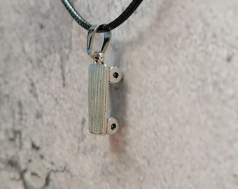 May include: A silver-colored pendant necklace with a rectangular design. The pendant is suspended from a black cord. The pendant has a textured surface and small, dark accents on each side. The background is a mottled gray.