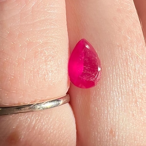 May include: A teardrop-shaped, vibrant pink gemstone. The gem is held between fingers, next to a thin silver ring. The stone's facets catch the light, displaying its polished surface. The background is dark, emphasizing the gem's color and shape.