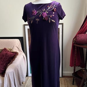 May include: A dark purple velvet dress with short sleeves and a floor-length skirt. The dress features floral embroidery in shades of purple, pink, and gold near the neckline. The dress is displayed on a mannequin.