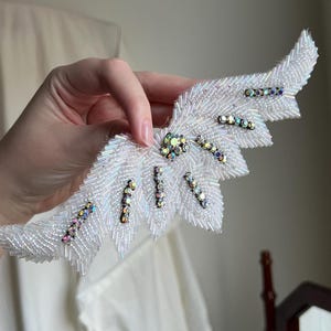 May include: A wing-shaped applique crafted from white iridescent seed beads, featuring a central cluster of clear crystals and several diagonal rows of multi-colored, reflective rhinestones embedded within the beaded structure.