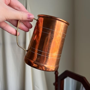 May include: A cylindrical copper mug with a polished metallic finish and a thin brass handle. The exterior surface features two sets of horizontal engraved lines encircling the body, providing a subtle decorative texture to the warm-toned metal.