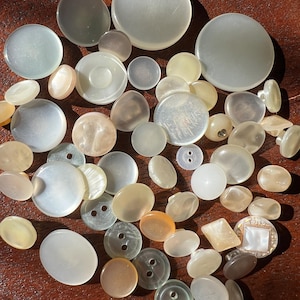 May include: A collection of various-sized, iridescent buttons in shades of white, cream, and pale yellow. Some buttons have two or four holes, while others are smooth and round. The buttons are scattered on a brown surface.