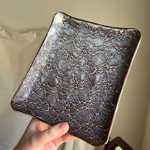May include: A rectangular ceramic plate with a floral lace pattern. The plate has a dark brown rim and a blue and white floral design. The plate is held in a hand.