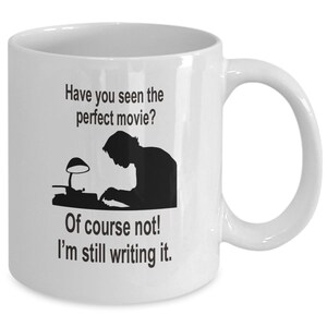 May include: White ceramic coffee mug with a black silhouette of a person writing on a typewriter. The text on the mug reads "Have you seen the perfect movie? Of course not! I'm still writing it."
